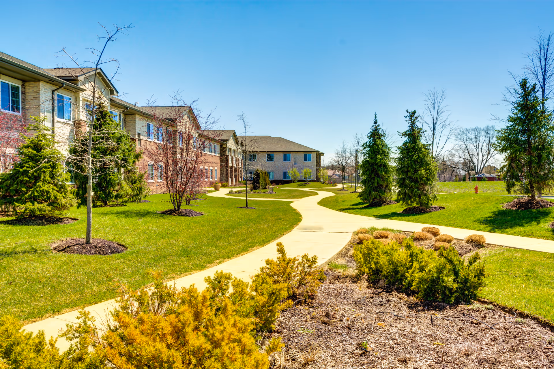 Curving paved walkway through landscaped lawns and trees in front of a two-story senior living building under a clear blue sky.