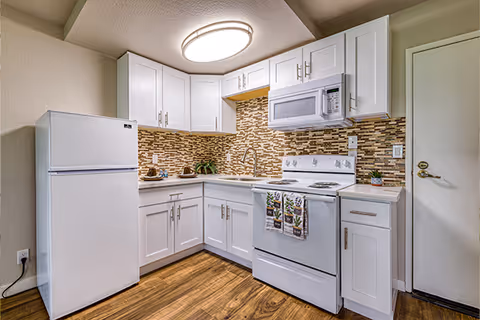 A compact kitchen with white cabinets, a white refrigerator, a white stove with oven, and a white microwave mounted above the stove. The backsplash features a mosaic tile pattern in shades of brown and beige. The kitchen has a wooden floor and a round ceiling light fixture. There is a door to the right side of the kitchen.