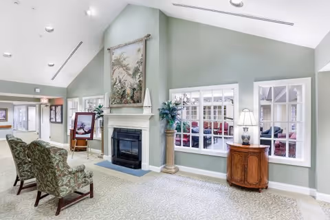 A bright and spacious senior living facility common area with high vaulted ceilings, light green walls, and patterned carpet. The room features a fireplace with a decorative tapestry above it, two upholstered armchairs, a wooden side table with a lamp, and large windows looking into another seating area with red sofas.