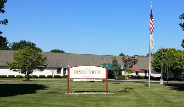 Front lawn and entrance of Devon House Senior Living with a sign, flagpole, and a single-story building in the background.