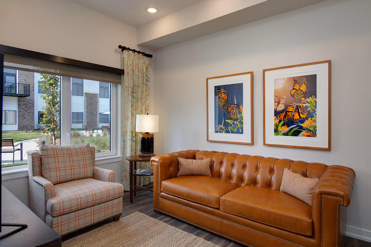 Cozy sitting area with a tufted leather sofa, plaid armchair, side table and lamp, and two framed butterfly prints on the wall.