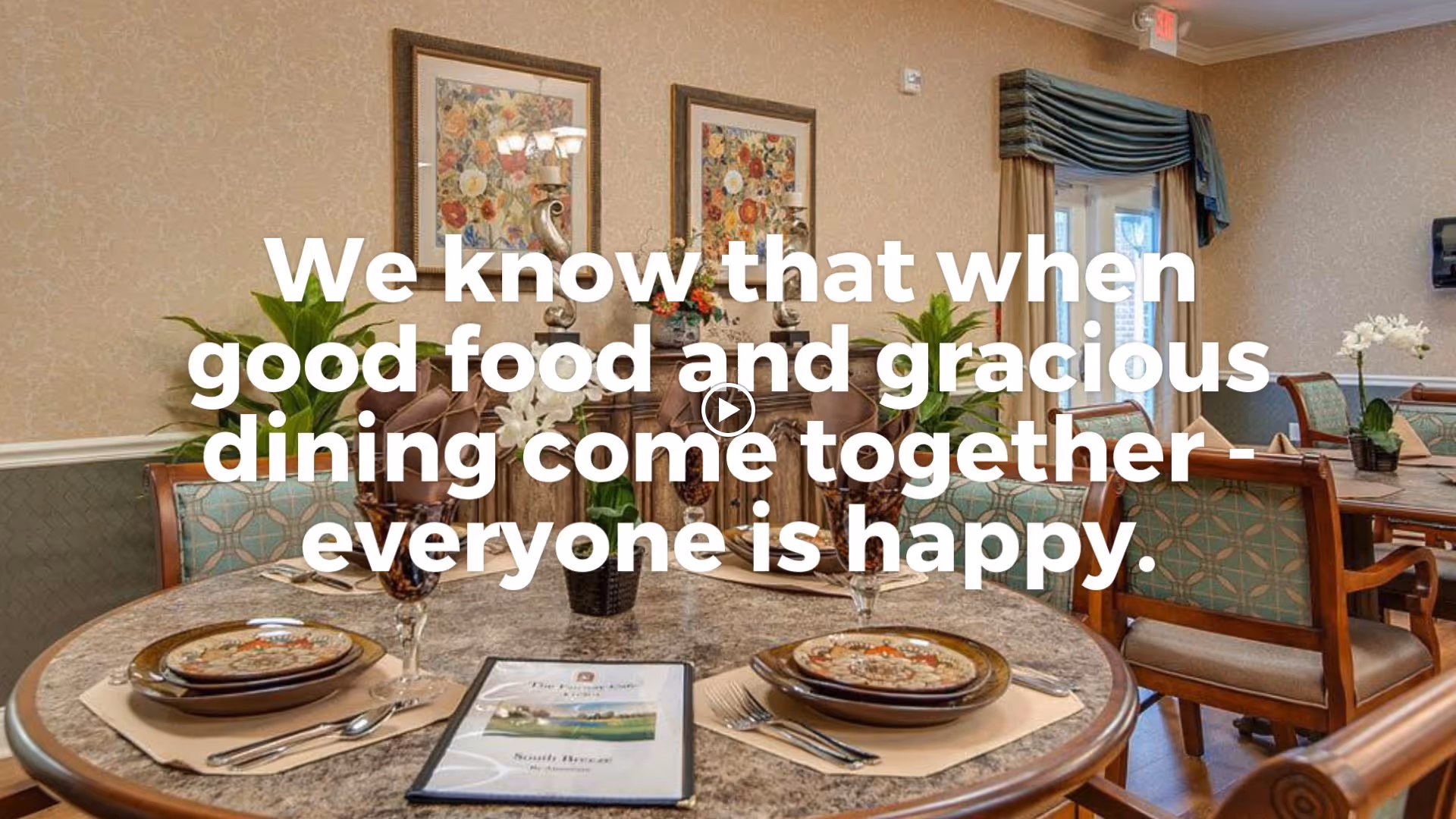 A warmly decorated dining room with round tables set for meals, featuring plates, glasses, and silverware. The room has floral artwork on the walls, plants, and large windows with curtains allowing natural light to enter.
