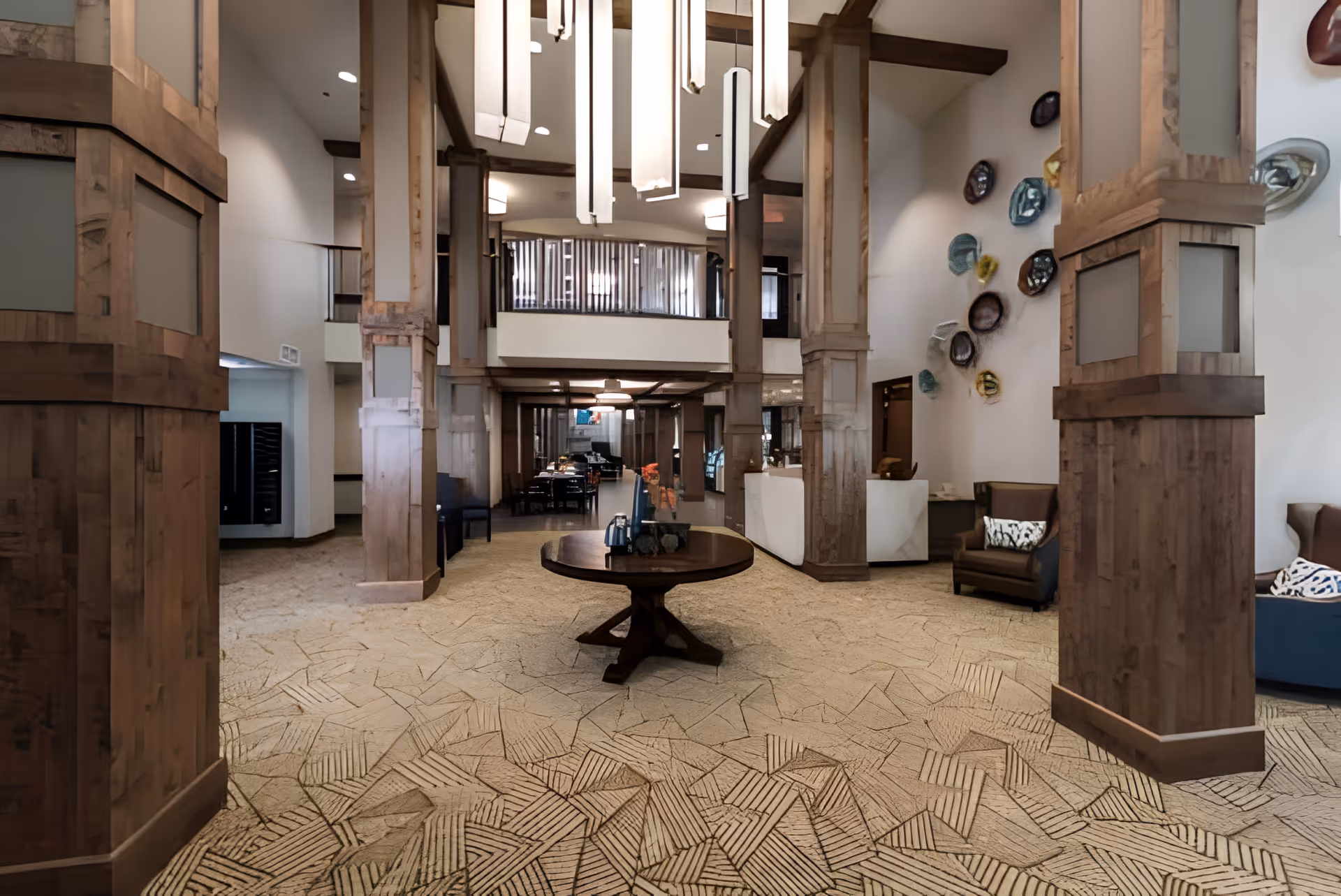 Spacious senior living lobby with wooden columns, seating areas, a central round table, and modern hanging lights.