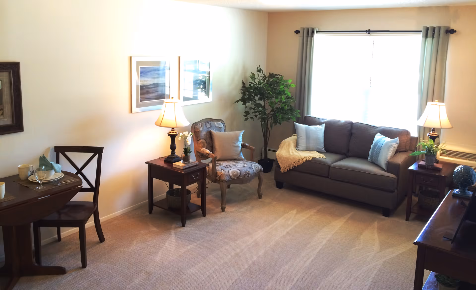 A cozy living room with a gray sofa adorned with three pillows and a beige throw blanket. Next to the sofa is a wooden side table with a lamp and a small plant. There is an upholstered armchair with a decorative pillow beside another wooden side table with a lamp and a small plant. A large window with gray curtains lets in natural light. On the left side, there is a small dining table set with two chairs, plates, cups, and napkins. The walls are decorated with framed artwork, and the floor is carpeted.
