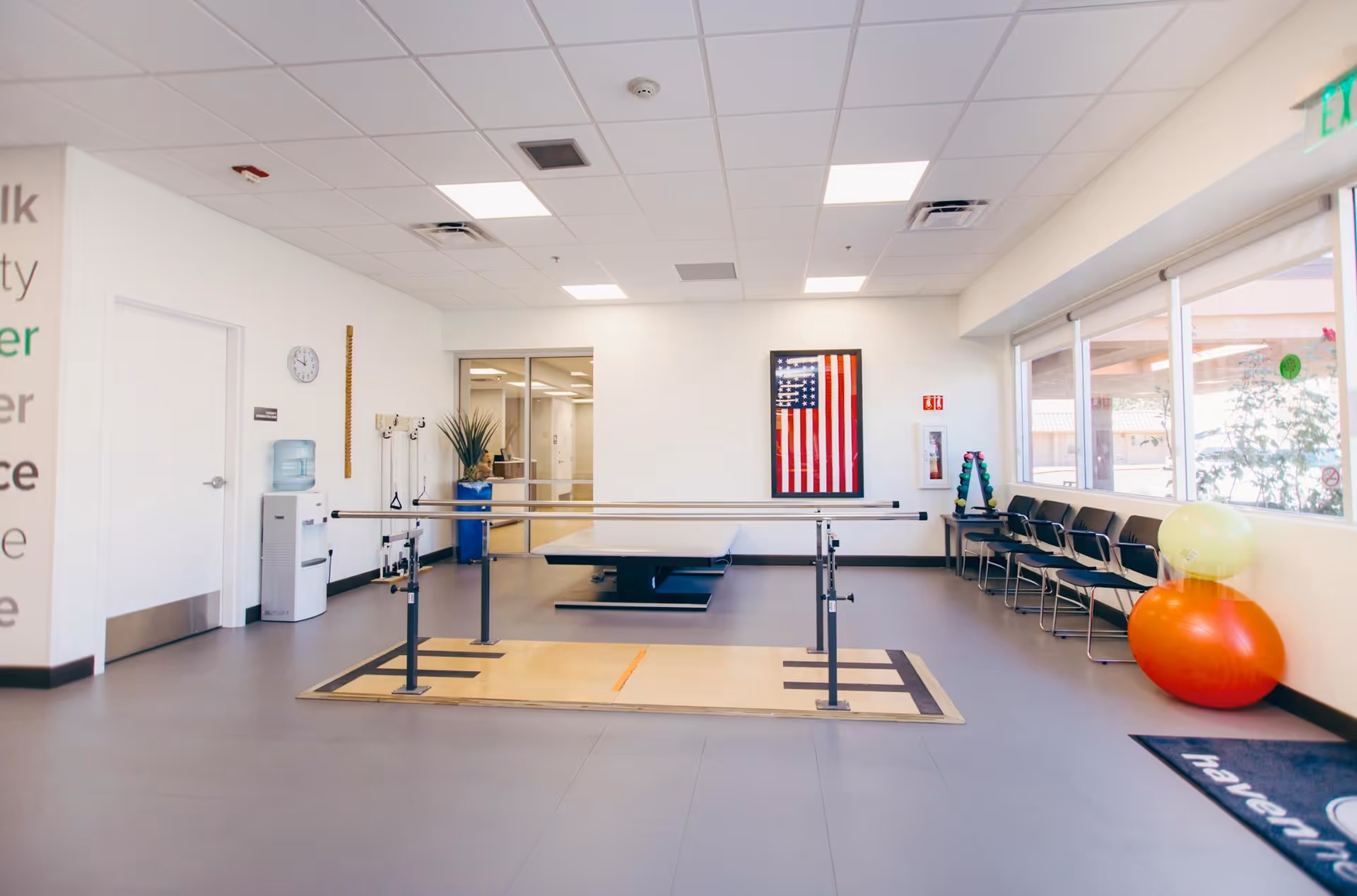 A spacious rehabilitation or physical therapy room with parallel bars for walking exercises, a therapy table in the background, chairs lined up along the window, exercise balls, and an American flag on the wall. The room has a water cooler, a clock, and a clean, bright interior with large windows letting in natural light.