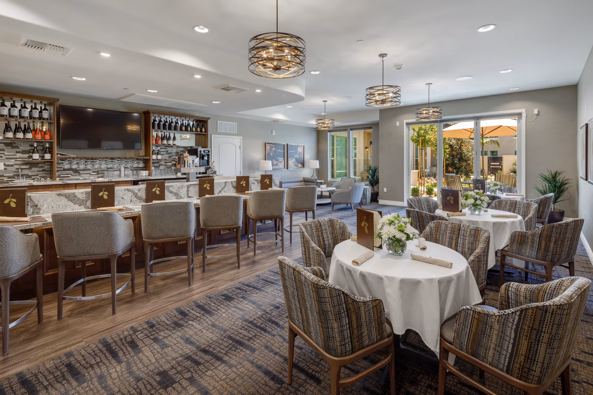 A spacious and well-lit dining area in a senior living facility featuring a bar with high chairs, tables with white tablecloths, floral centerpieces, and comfortable upholstered chairs. Large windows and glass doors provide a view of an outdoor patio with umbrellas and greenery. Modern light fixtures hang from the ceiling.