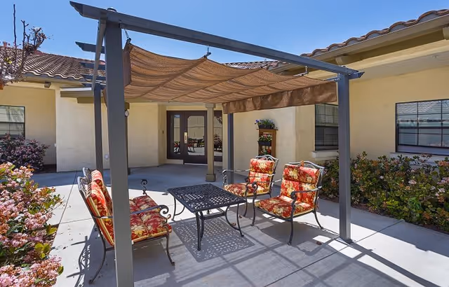 Outdoor shaded courtyard with a pergola, metal table and cushioned chairs outside the facility entrance.