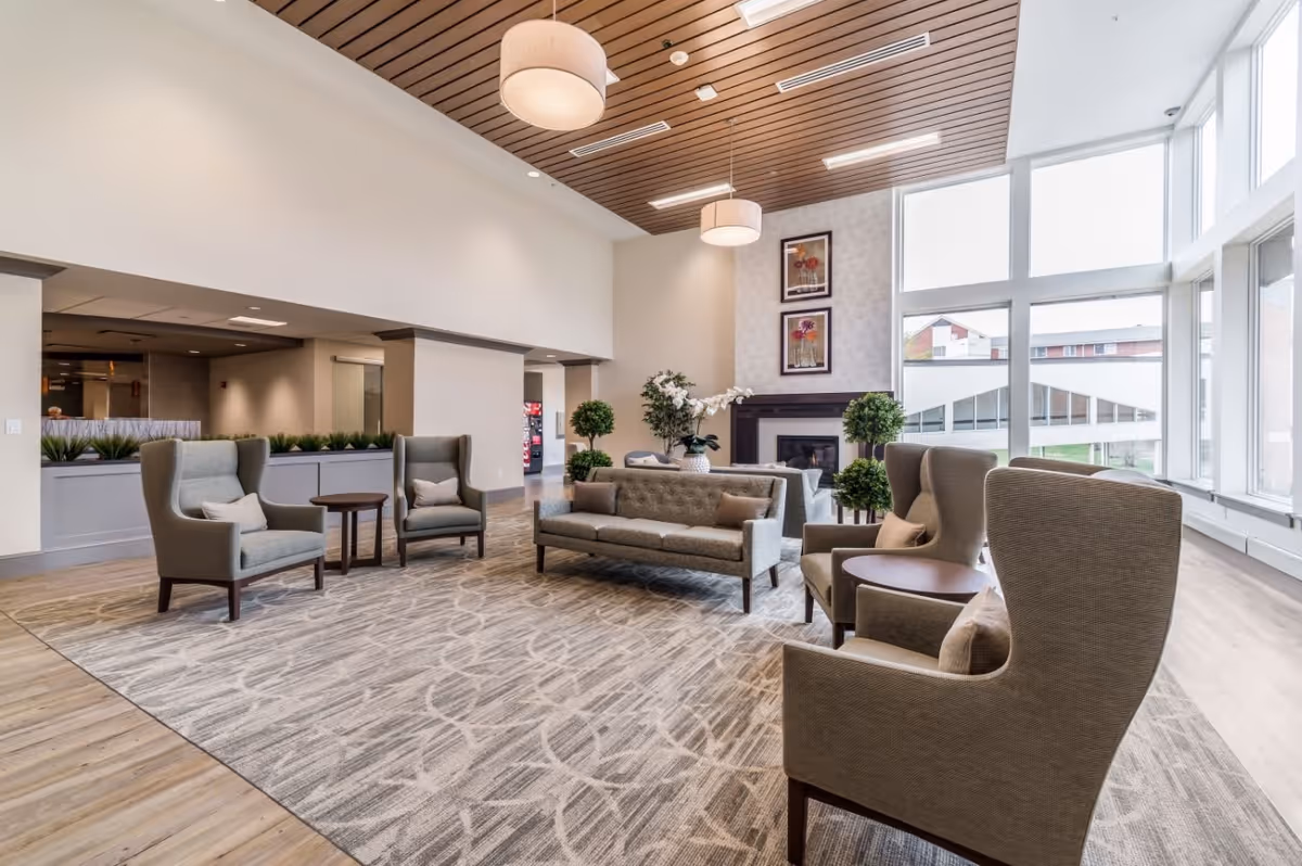 A spacious senior living facility lounge area with modern furniture including a gray sofa, four armchairs with cushions, and two round wooden side tables. The room features a patterned carpet, wood-paneled ceiling with hanging light fixtures, large windows letting in natural light, and a fireplace with two framed floral artworks above it. There are also several potted plants adding greenery to the space.