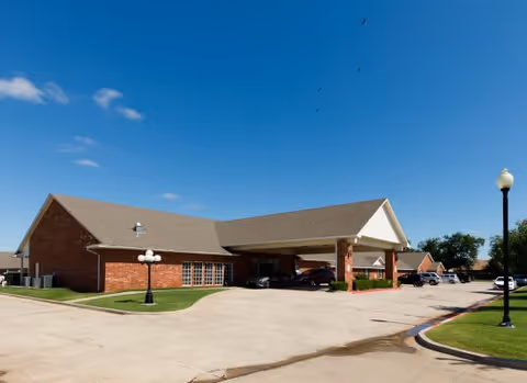 Exterior view of a single-story brick building with a covered entrance and several parked cars under a clear blue sky. The building is surrounded by a paved driveway and green grass with street lamps.
