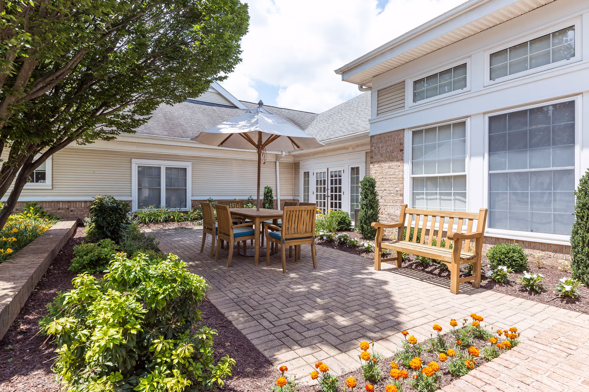 Outdoor patio area at Copper Ridge featuring a wooden table with six chairs under a large white umbrella, surrounded by green shrubs, flowers, and trees. The patio is paved with bricks and adjacent to a building with large windows and white trim.