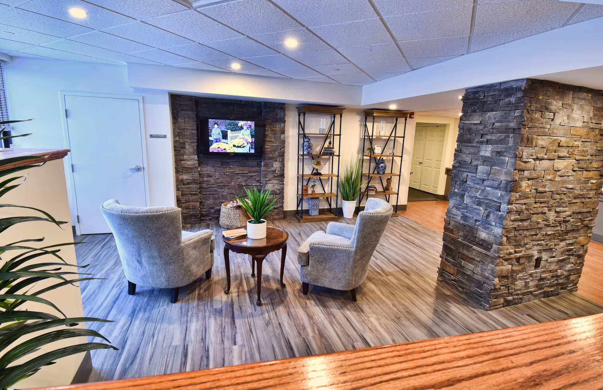 Cozy seating area with two upholstered chairs around a small wooden table facing a wall-mounted TV, with stone pillars, shelving units, plants, and wood-look floors.