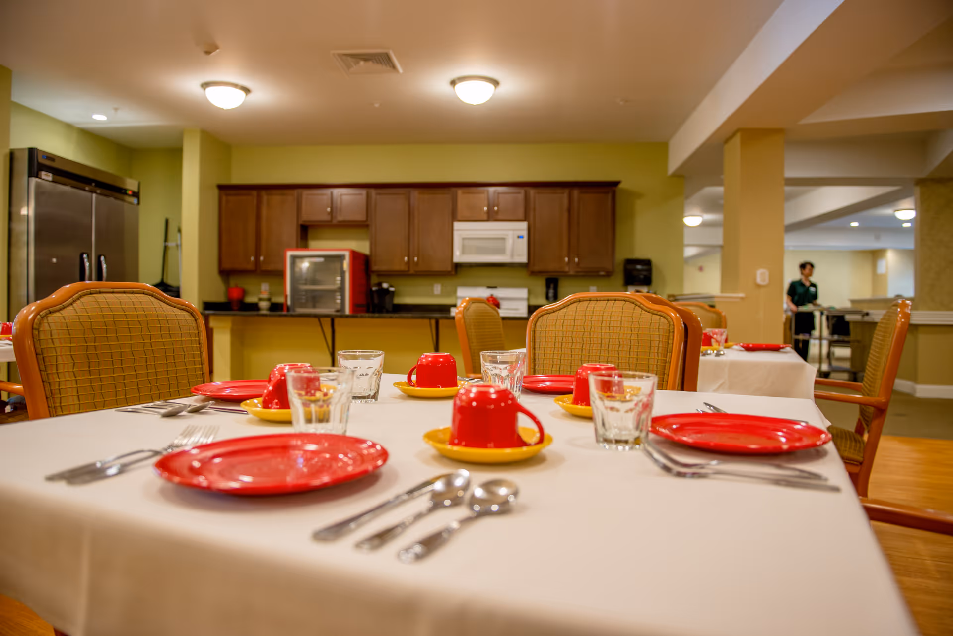 Dining area in Linda Manor Assisted Living with tables set with red plates, red cups on yellow saucers, glasses, and silverware. The background shows a kitchen area with wooden cabinets, a microwave, and a refrigerator. A staff member is seen in the distance near another table.
