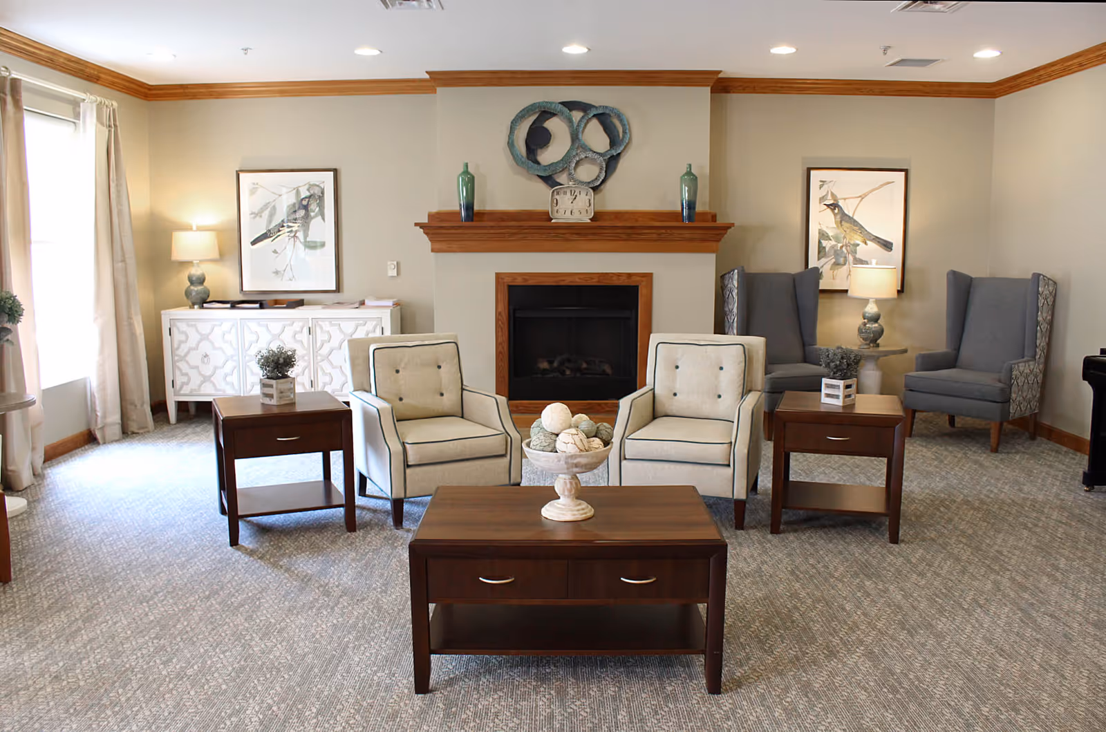 A cozy living room with two beige armchairs and two gray wingback chairs arranged around a wooden coffee table and two side tables. The room features a fireplace with a wooden mantle decorated with two green vases, a clock, and a circular wall art piece. There are framed bird prints on the walls, a white cabinet, and a window with beige curtains letting in natural light.