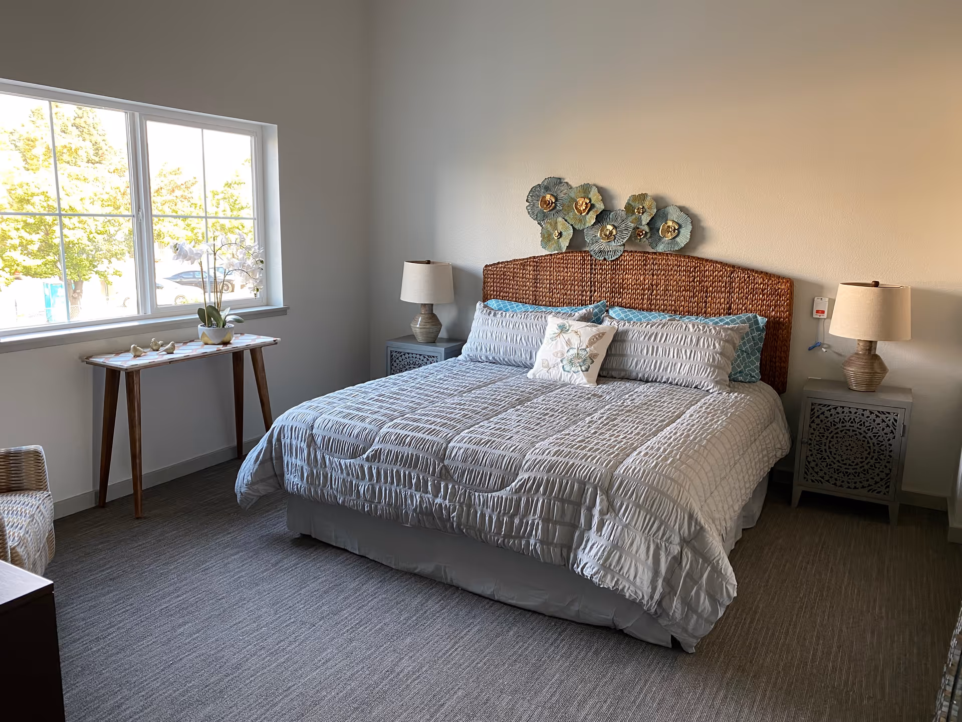 A neatly made bed with a woven headboard and decorative pillows in a bright bedroom. There are two bedside tables with matching lamps on each side of the bed. A small wooden table with decorative items and a potted orchid is placed near a large window letting in natural light. The room has light-colored walls and carpeted flooring.