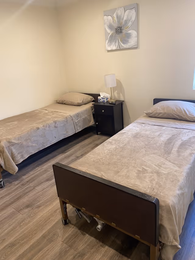 A simple bedroom with two single beds covered in beige blankets and pillows. Between the beds is a small black nightstand with a white lamp, some tissues, and a few small items. Above the nightstand hangs a painting of a large white flower. The room has light-colored walls and wood flooring.
