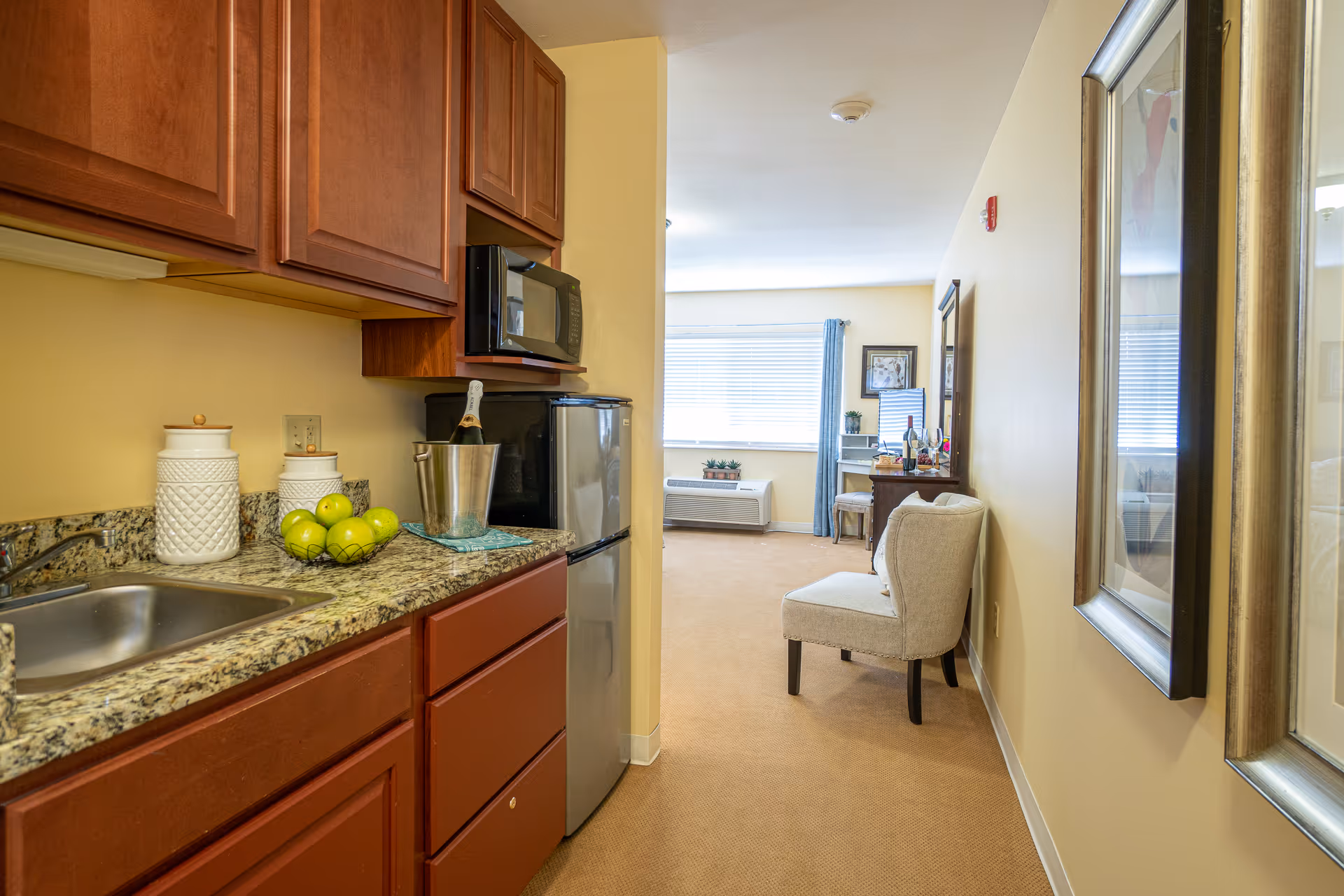 View of a small kitchen area with wooden cabinets, granite countertop, a sink, a microwave, and a mini refrigerator. On the counter, there are three white canisters, a bowl of green apples, and a champagne bottle in an ice bucket. The kitchen opens into a living space with a beige carpet, a cushioned chair against the wall, a window with blue curtains, and a desk with a mirror and decorative items.