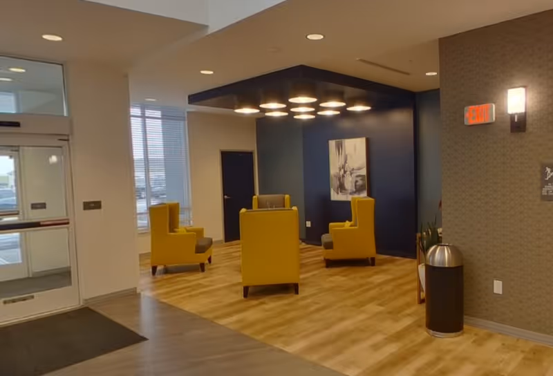 Interior view of a senior living facility lounge area with four yellow armchairs arranged in a square on a wood floor. A dark blue accent wall features a modern abstract painting and overhead circular lights. There is a glass door entrance on the left and an exit sign on the right wall above a trash can.
