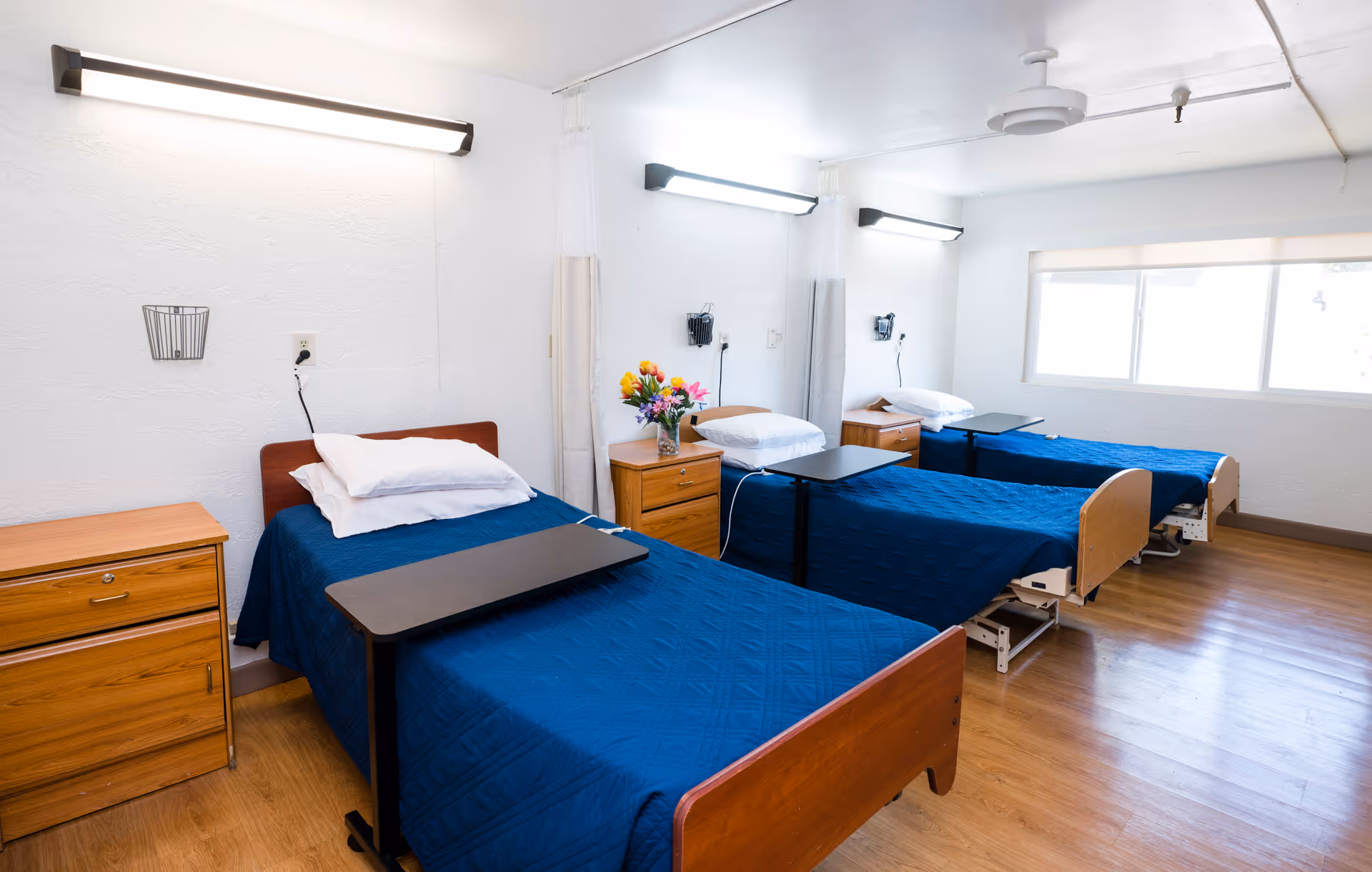 A bright shared patient room with three hospital beds covered in blue quilts, bedside tables, and overbed trays.