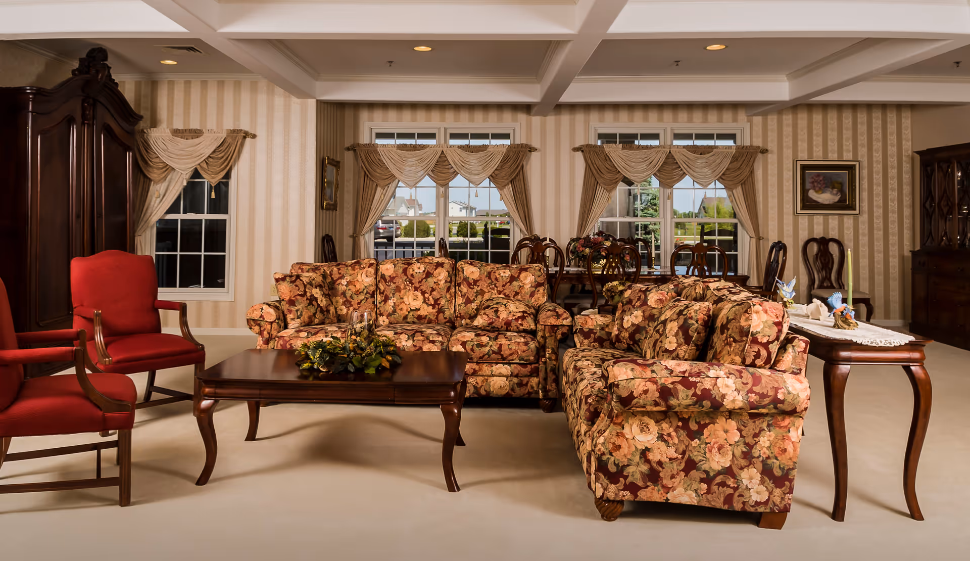 A traditional living room with floral patterned sofas, two red armchairs, a wooden coffee table with a floral centerpiece, and a wooden side table with decorative items. The room has beige striped wallpaper, three windows with beige drapes, and a dining table with chairs in the background.