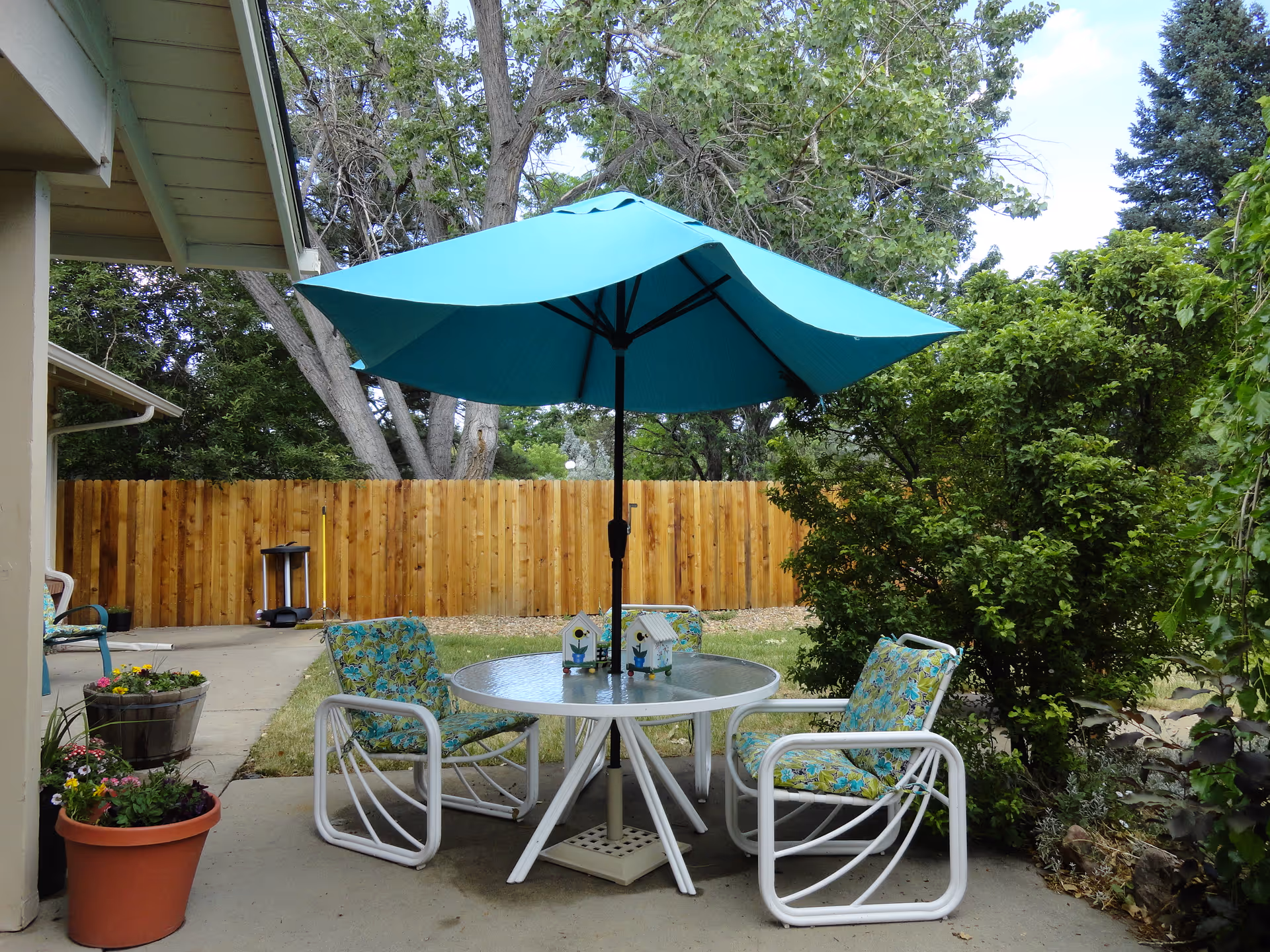 Outdoor patio area with a round glass table, three white metal chairs with blue floral cushions, and a large blue umbrella providing shade. There are potted plants with flowers on the concrete patio, a wooden fence in the background, and green trees and bushes surrounding the area.